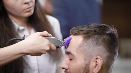 Obraz na płótnie Canvas Close up of professional female hairstylist dressed in casual clothes is serving client in barbershop using modern trimmer. Young man is getting trendy haircut sitting on chair closing his eyes.