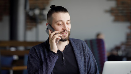 A young attractive hipster in a cafe, with a modern interior. A man is talking on the phone with a friend or colleague. The guy break, he is in a good mood.