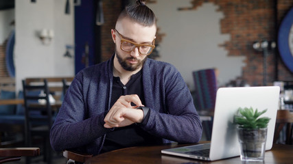 Close up of bearded hipster sitting in restaurant, looking and touching smartwatch. Male designer is waiting for client at the table in cafeteria with laptop and modern wristwatch on his hand.