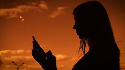 Silhouette of an adult girl against the sky, woman with long hair prints a message on a mobile phone. It uses a gadget to communicate with friends
