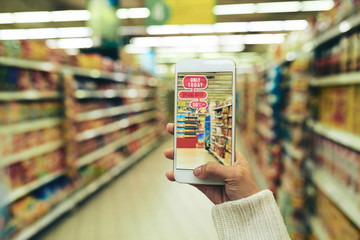 Close-up shot of young woman doing shopping at hypermarket with help of augmented reality app, blurred background