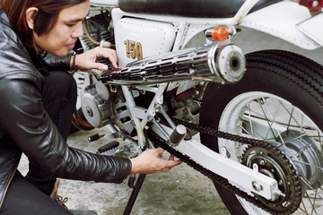 Confident Asian man wearing black leather jacket sitting on haunches while adjusting chain of vintage motorcycle
