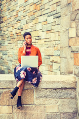 Fototapeta premium Young African American Woman working on laptop computer, talking on cell phone outside in New York, wearing orange red jacket, flower patterned skit, black boot shoes, sitting by stone wall on campus.