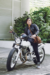 Fototapeta premium Full length portrait of brutal Asian biker wearing black leather jacket posing for photography while sitting on vintage motorcycle outdoors