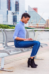 Young African American college student studying, working on laptop computer outside in New York, wearing striped shirt, jeans, boot shoes, sitting on metal chair at park, looking down, reading, typing