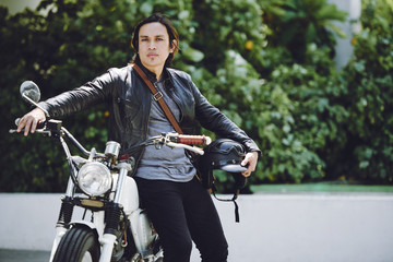 Brutal Asian man looking at camera and leaning on vintage motorcycle while taking short break from...