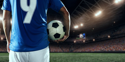 Soccer player holds a football ball on a professional stadium. Player wears blue unbranded uniform.