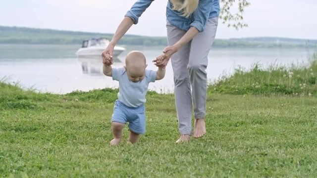 Unrecognizable mother holding hands of cute baby boy walking on green grass in park with lake