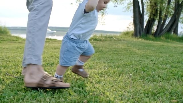 Tracking of unrecognizable mother holding hands of adorable toddler boy walking on green grass in park with lake on summer day