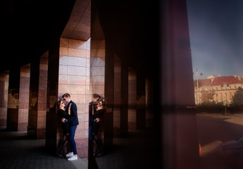 Man in black jacket and jeans leans to a marble wall hugging his girlfriend