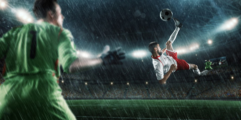 Soccer player scores the ball into the goal on professional rainy stadium. The goalkeeper protects...