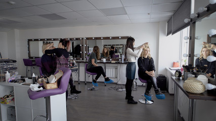 Fototapeta premium Professional hair stylist and makeup artist in their large studio preparing models before going on the podium for the show. Beautiful girl admires itself in a mirror and then looks in the smartphone.