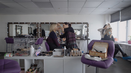 Obraz premium Beauty shop. The master in a make-up trains three girlfriends to final at university. The girl applies with a shadow brush on top. The makeup artist professionally does a make-up.