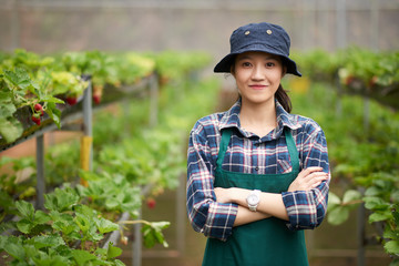 Waist-up portrait of smiling Asian gardener wearing apron and bucket hat standing at modern spacious greenhouse with arms crossed and looking at camera