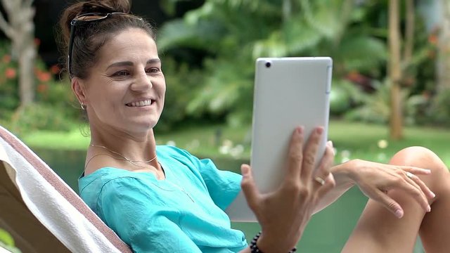 Beautiful woman having a videocall on tablet while sitting next to the swimming pool, steadycam shot
