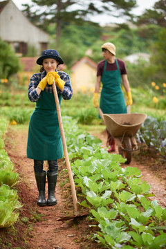 Full length portrait of beautiful Asian woman wearing gumboots and apron posing for photography with wide smile while leaning on hoe, spacious vegetable garden on background