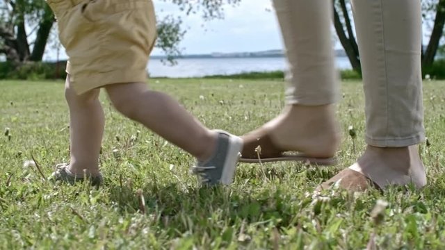 Tracking with low-section of legs of unrecognizable woman and toddler walking on green grass in park