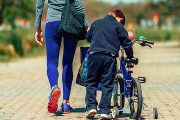Child carries his bike while walking through a park with his mother