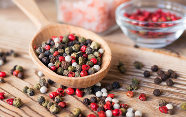 Mix of peppercorns in wooden spoon on rustic background with pink salt in glass jar. Red, black, green and white dry pepper in seeds.