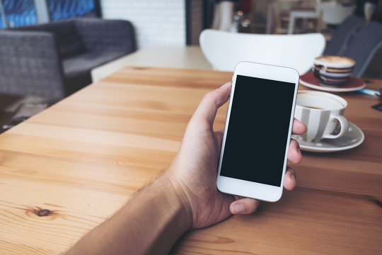 Mockup image of a man's hands holding white mobile phone with blank black screen and white coffee cups in modern loft cafe