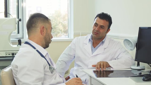 Two doctors are sitting in the office. They are talking. One and the doctors said something funny and both laugh. Doctors look very friendly and nice