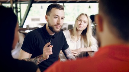 An adult and bearded man with tattoos on his hands tells a funny story, a company of friends is resting and laughing at the bar