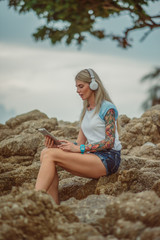 Beautiful blonde woman sitting on a rock by the sea and listening to music with your tablet. hands with tattoos. Style summer vacation. Modern girl