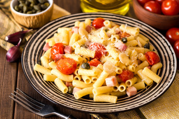Pasta with bacon, garlic, tomatoes, capers and parmesan cheese in plate on dark wooden background.