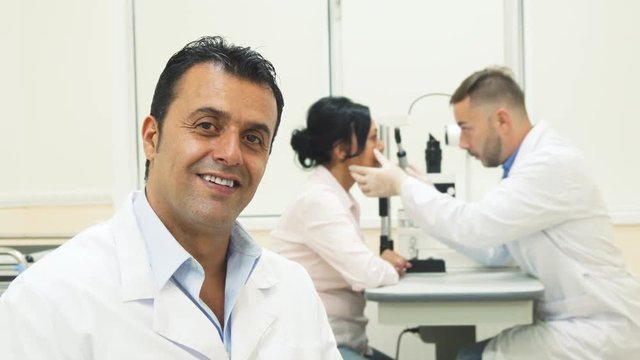 The doctor is sitting in the foreground. He smiles and looks very friendly. In the background is another doctor. He makes the diagnosis of the patients eyes