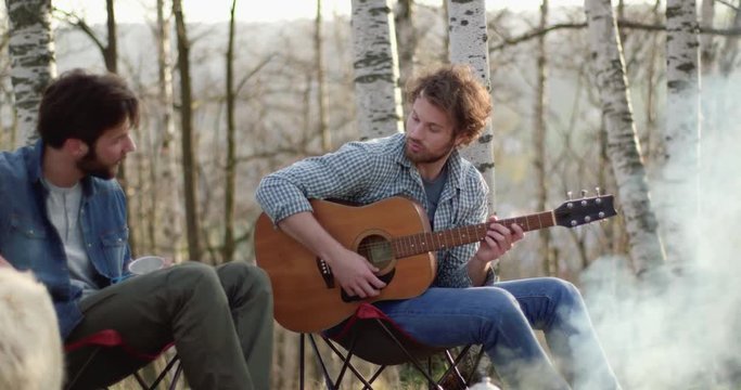 Man playing guitar and friend stroking dog together around fire place.real friends people outdoor camping tent vacation in autumn trip.Fall sunny day in nature, togetherness and friendship. 4k video