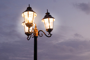 Old fashioned street lights with three lamps set on a pole shot against a cloudy sky. The dusk light and orange glow of the lamps gives this a perfect old time feel
