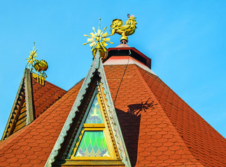 A pointed roof with a cock on top and a triangular stained-glass window against the blue sky
