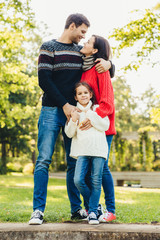 Fototapeta premium Young happy family of three stand together outdoors, embrace, have good relationships. Couple in love look at each other, hug their little daughter. Happiness and harmony in family life.