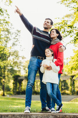 Fototapeta premium Vertical portrait of family of three members stand close to each other, embrace, have walk across autumn park, notice squirrels on tree. Friendly father, mother and daughter have good relationship