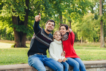 Fototapeta na wymiar Father and mother sit between their daughter against trees or nature background, show something to her, indicate with fore fingers. Smiling parents show bird to little beautiful girl. Parenthood