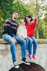 Fototapeta na wymiar Carefree restful woman, man and small cute girl sits on bridge outdoors, admire sunrise. Affectionate mother shows to her little daughter beauty of nature. Family enjoys spending free time in park