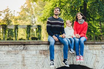 Horizontal portrait of adorable child sits between her mother and father, enjoy beaitiful landscapes from top of ancient bridge, breath fresh air, look into distance. Family and relationship concept