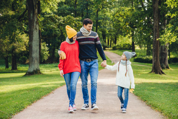 Lovely couple embraces each other, looks at their small good looking daughter, walks across green park. Little adorable girl keeps father`s hand, has pleasant conversation with her parents