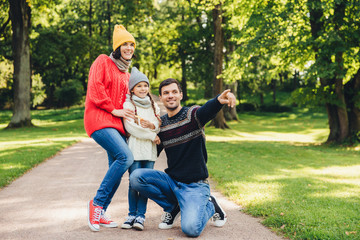 Fototapeta premium Horizontal portrait of family with good relaionship stand close to each other, admire landscapes or beutiful nature. Happy father shows to her daughter big old tree in park, indicates with fore finger