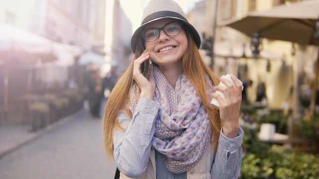 Joyful caucasian girl, in striped hat and glasses, walking backward with coffee and mobile phone, sunny day, outdoor slowmotion