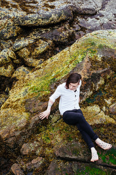 Transgender, gender fluid person hanging out on algae covered rocks at the beach.