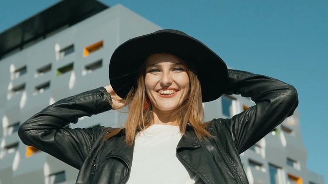 Happy caucasian girl, in fashionable black hat and leather jacket, posing before modern building, slowmotion
