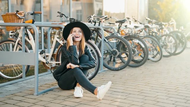 Cheerful caucasian girl, in black hat and leather jacket, sitting before bikes and talking on the phone, outdoor slowmotion in sunset time
