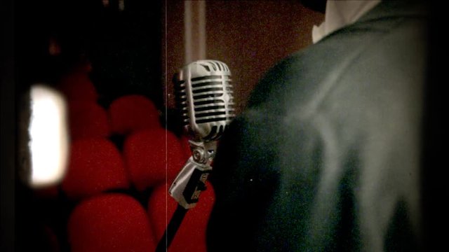 Fake 8mm amateur film: a singer in front of a retro vintage shiny microphone on the stage of a theater or cinema. Red seats in the background.
