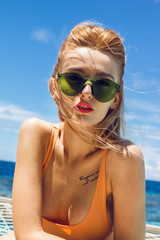 Beautiful young blonde girl lying on a networked terrace over the open ocean. She has a stylish green sunglasses that fits well with her orange bikini and red lipstick.