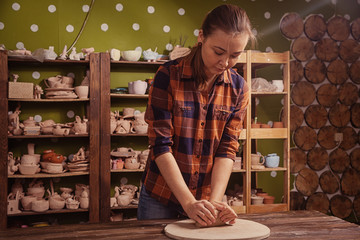 A young woman in a plaid shirt and jeans kneads the clay.  before working on the pottery wheel of a plate on a wooden circle in the workshop, in the background, racks with clay pots
