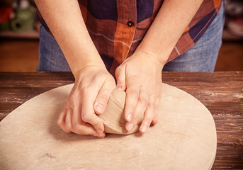 A young woman in a plaid shirt and jeans kneads the clay with her hands before working on the pottery circle of a plate on a wooden circle in the workshop