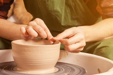Fototapeta na wymiar Close-up A woman potter in a plaid shirt and green apron beautifully sculpts a deep bowl of brown clay and cuts off excess clay on a potter's wheel in a beautiful workshop