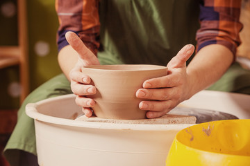 A female potter in a green apron and plaid shirt sculpts clay vase from the clay on a potter's wheel in the workshop