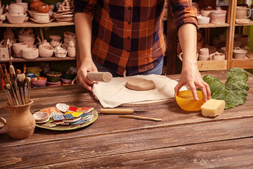 Woman  potter in a plaid shirt and jeans rolls out clay to make a plate on a wooden table in the workshop, on the table there are clay fakes, in the background, the shelves are in clay cups and vases